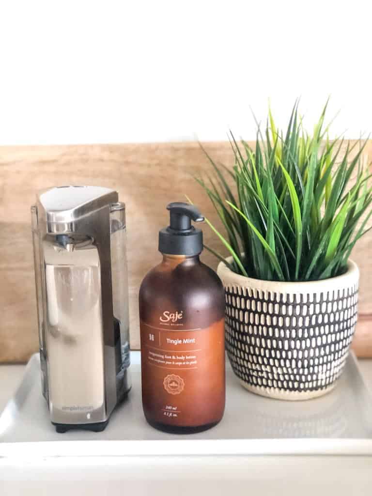 Signed Samantha's COVID-friendly must haves includes a touchless soap dispenser which is pictured on a white tray with saje lotion, and a plant.