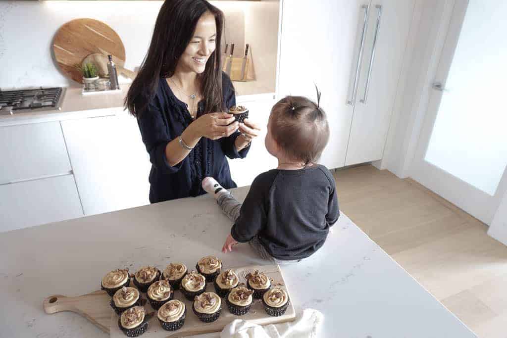 Signed Samantha is feeding her daughter, who is sitting on the kitchen counter, gluten-free chocolate cupcakes. A whole cutting board filled with them is also sitting on the counter.