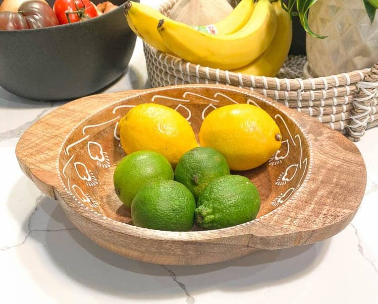 one wood bowl with lemons and limes, one black bowl with tomatoes and onions, and one wicker basket with bananas is pictured for Signed Samantha's post on how to style kitchen countertops