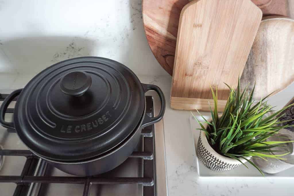 Signed Samantha's splurge worthy kitchen must haves include a matte liquorice colored le creuset dutch oven pictured on a gas stove, next to a countertop with a plant, salt and pepper shakers, and wooden cutting boards leaning against the wall.