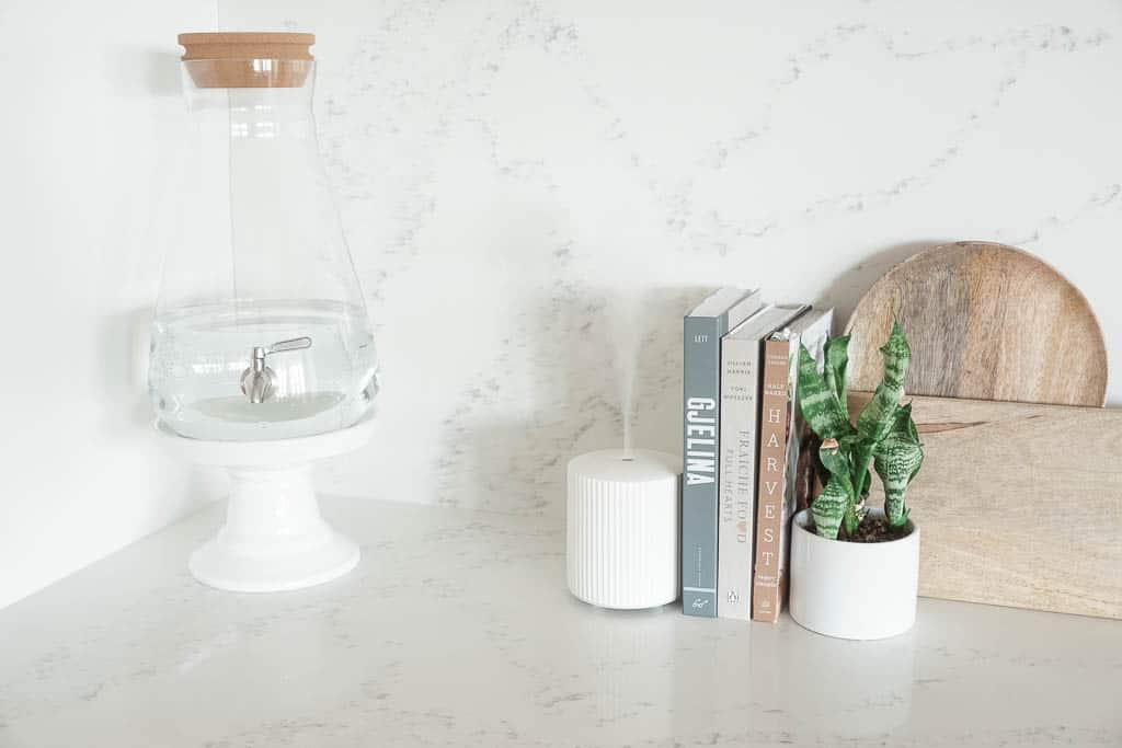 Signed Samantha's splurge worthy kitchen must haves include a huge glass carafe with a cork lid sitting on a ceramic base. Also pictured next to the ceramic base is a white diffuser, three cookbooks and a plant in front of two wooden cutting boards.