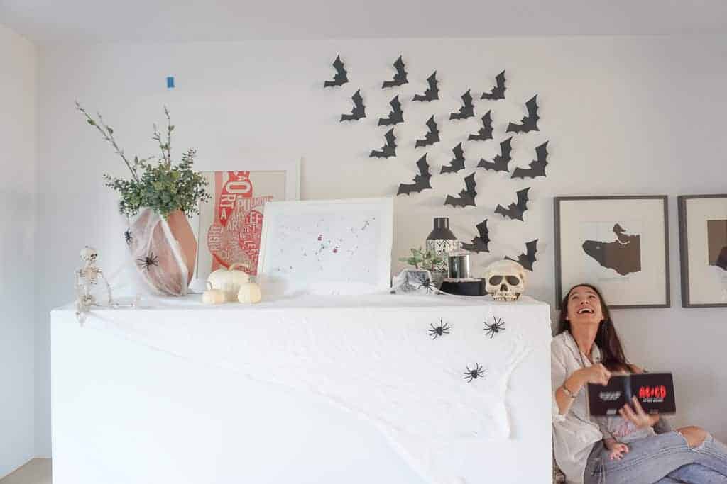 Signed Samantha sharing her D.I.Y Halloween Bat Decor - that's so cheap and so easy. There are many bats on the wall above her fireplace that look like they are flying away and she is looking up at them while her daughter reads a book on her lap.