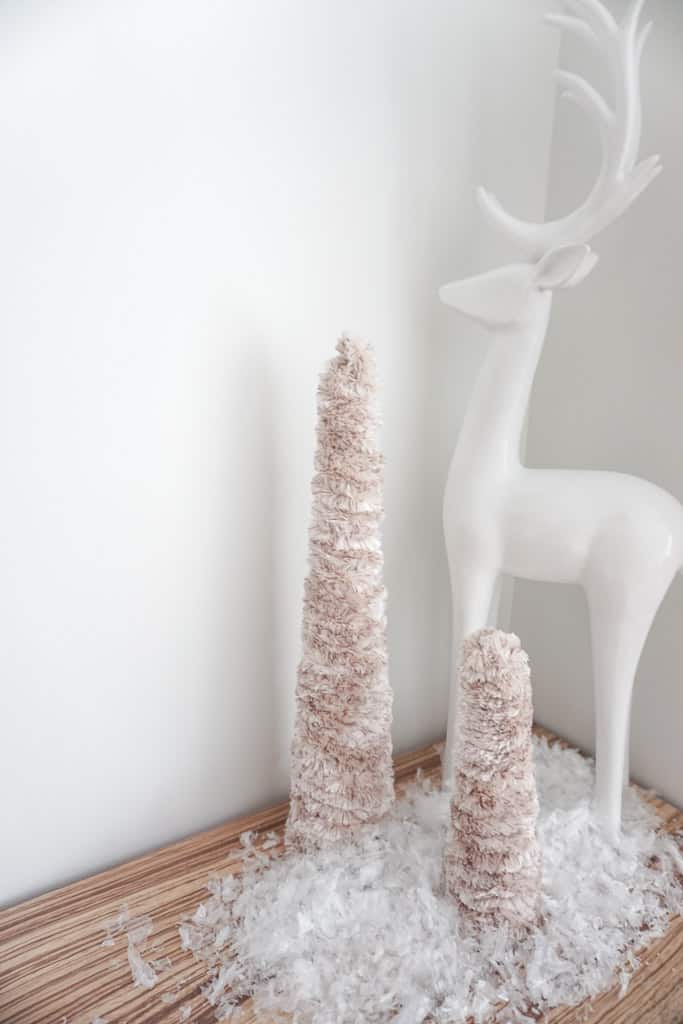 D.I.Y mini christmas trees and a white reindeer pictured on a wood table with faux snow in front of it.