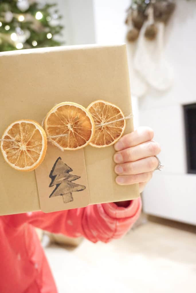 Signed Samantha holding up a holiday gift showing her D.I.Y holiday gift tags. The gift is wrapped in craft paper with dired oranges.