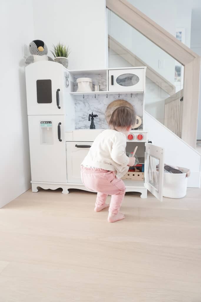Signed Samantha Potter sharing her super simple way to do a kids kitchen makeover with her daughter, Sloane's kitchen. Sloane is standing in front of the kids kitchen that has red knobs, a faux marble backsplash, and black handles.