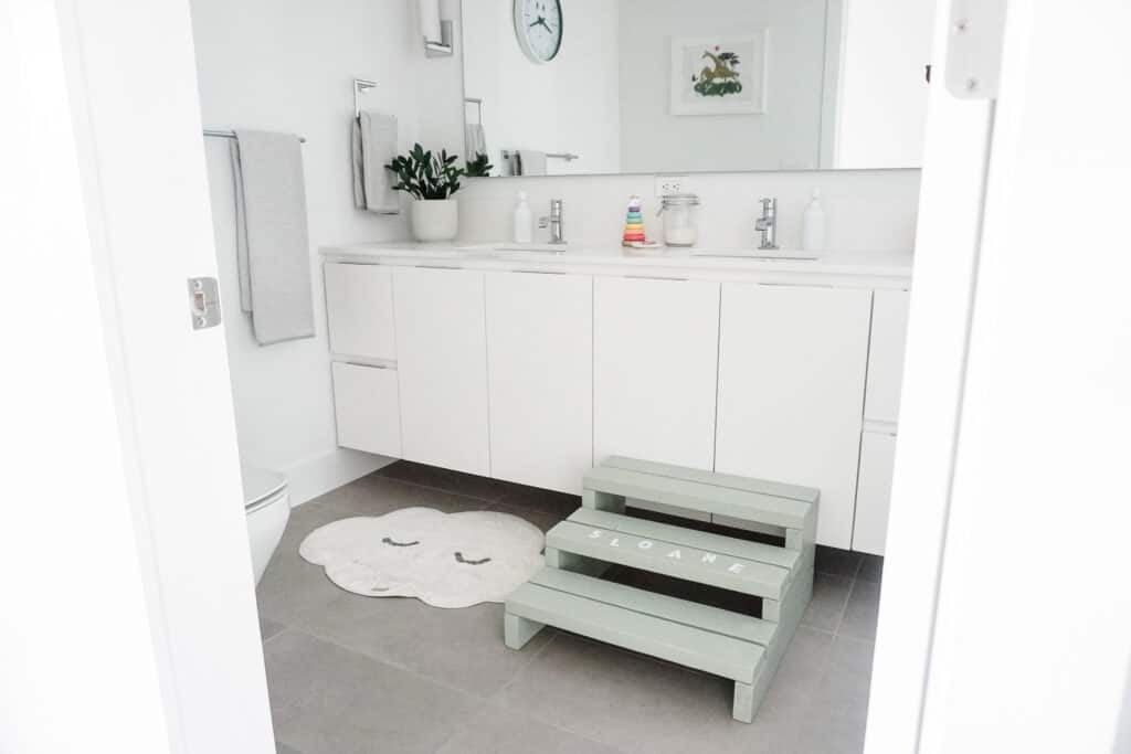 A sage green step stool for kids with Sloane's name on in it in white paint. Signed Samantha teaches you how to make your own step stool for kids. The stool is in a bathroom with two sinks and white cupboards and a big mirror.