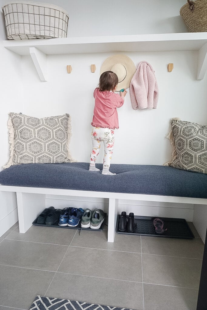 DIY Bench Cushion Cover in Samantha's mudroom - it is a blue thicker fabric material with throw pillows and a toddler jacket and hat on the hooks. Sloane is standing on the bench cushion playing with the hat