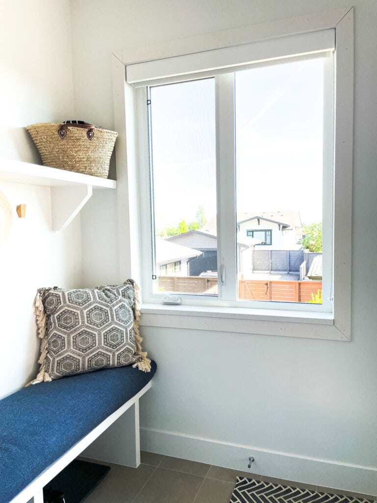 Sharing Samantha Potter's DIY Window Trim around the window in her mudroom. It is unpainted and not filled and sanded, yet. Next to a bench with a blue cushion on it and a decorative pillow.