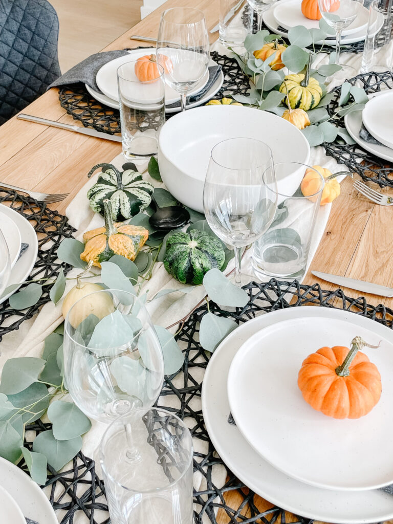 Setting the table for thanksgiving in Signed Samantha's house includes a runner down the middle of the table, black placemats, white dishwear, pumpkins, eucyalptus, and wine galsses