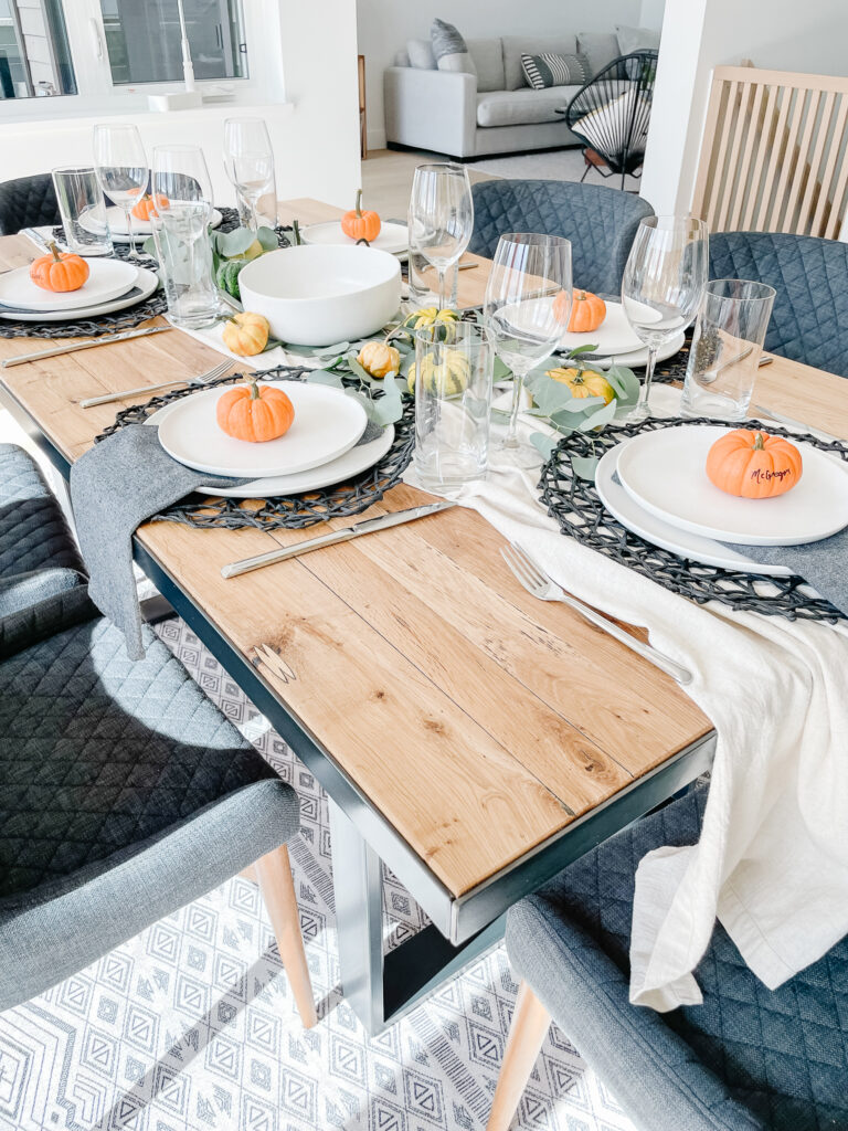 Setting the table for thanksgiving in Signed Samantha's house includes a runner down the middle of the table, black placemats, white dishwear, pumpkins, eucyalptus, and wine galsses