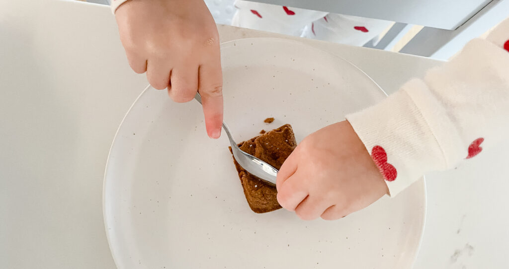 A toddler cutting up a piece of gluten-free brownie to eat.