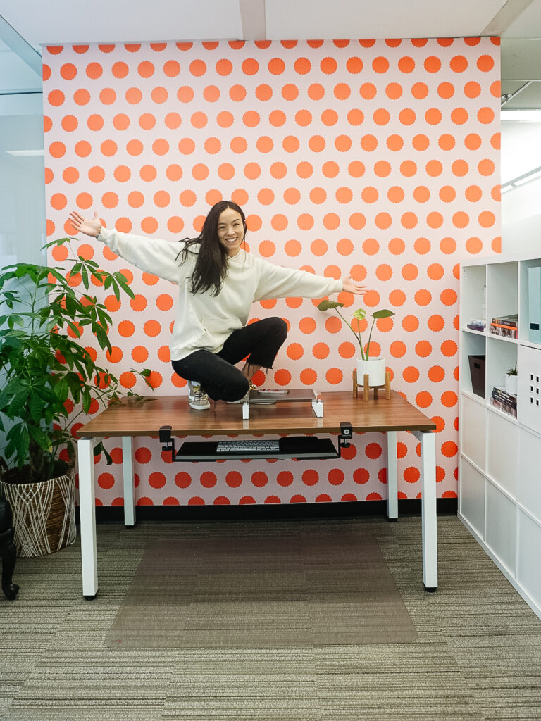 Otto studio's peel and stick wallpaper in polka pop - tangerine is on the wall with a desk and laptop in front. Signed Samantha is sitting on the desk in front