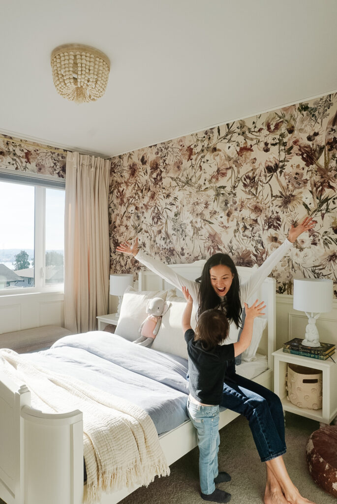 things to consider when costing a DIY project - A big girl room with floral wallpaper, chair rail, box moulding, a white bed frame, blue duvet cover, end tables, and a pouf on the ground plus signed samantha sitting on the bed and her daughter in front of her with her arms up