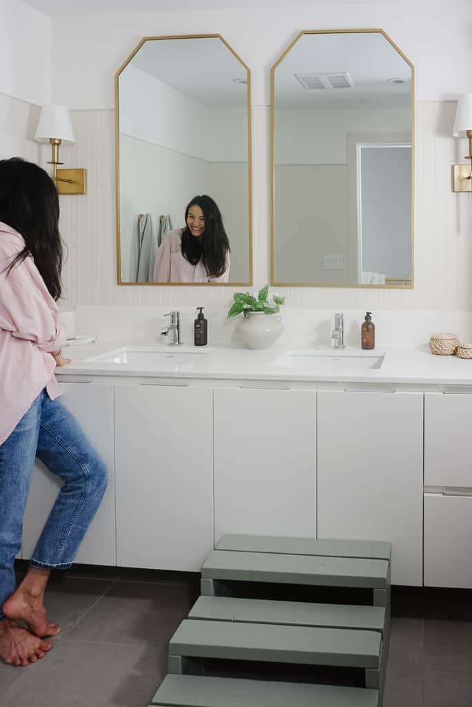 builder grade bathroom gets an upgrade with two gold mirrors, gold sconces, beadboard on the walls with a creamy white. Samantha is standing at the sink.