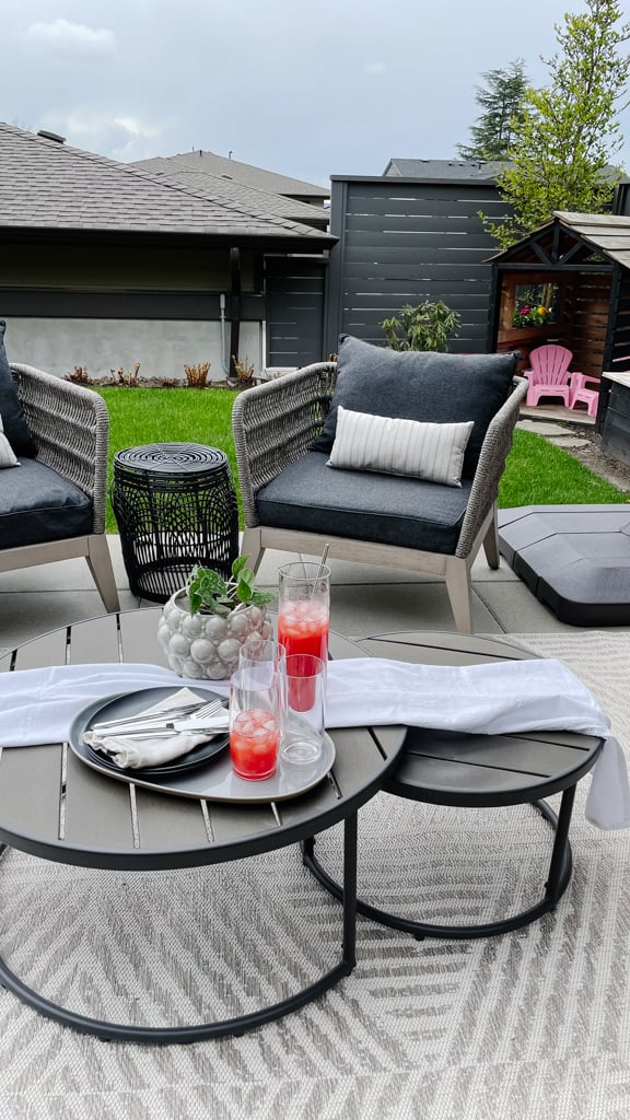 Outdoor patio oasis set up with club chairs, an inviting table setting with a red drink and cups, and green plants.
